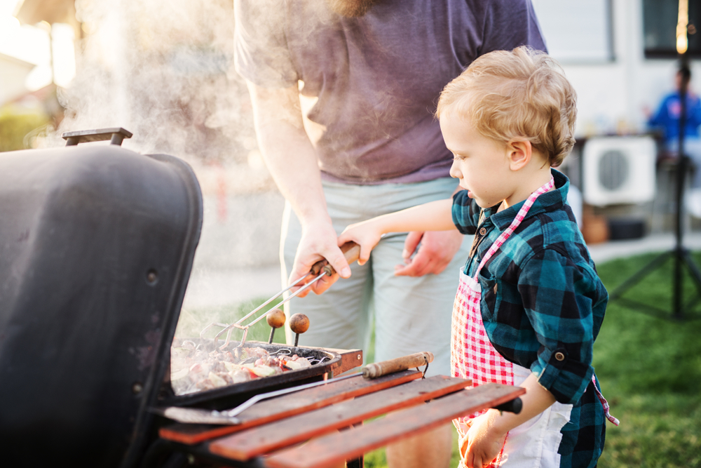 little boy is holding apron and learning how to make barbeque with parents help.