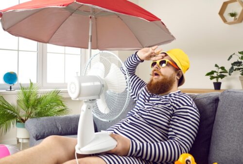 bearded man in swimsuit sitting at home, suffering from crazy summer heat, wiping sweat off forehead, holding electric fan, wishing for heatwave to stop and fresh breeze to blow
