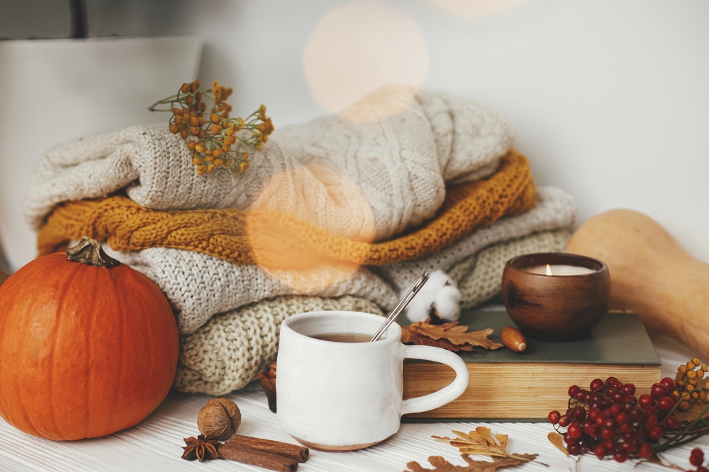 Pumpkin, cup of tea, cozy sweaters, autumn leaves, burning candle and vintage book in golden lights bokeh on white wooden background in room.