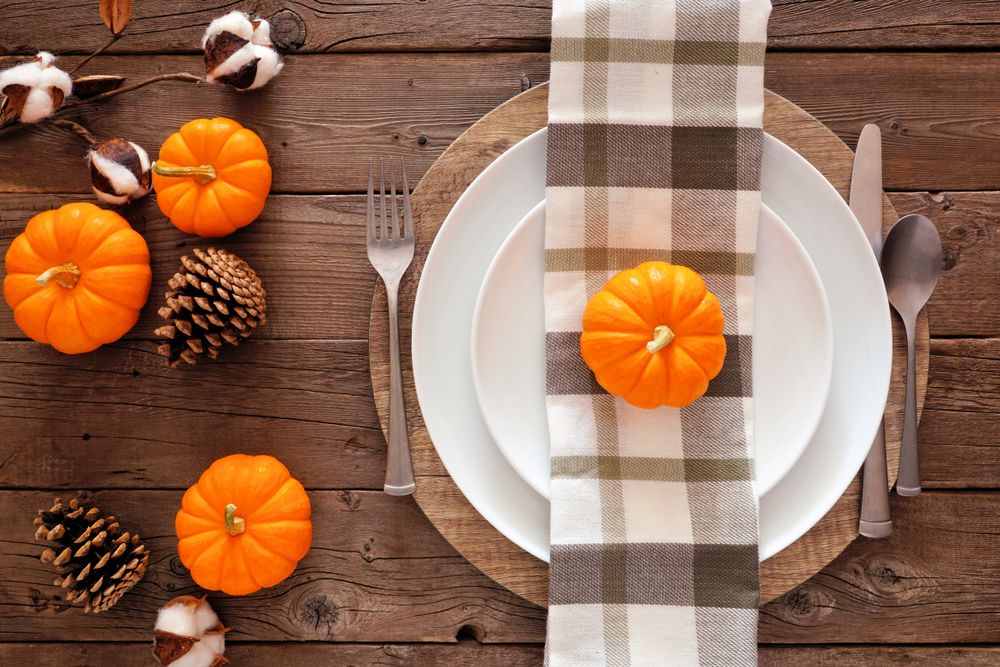 Autumn harvest or thanksgiving dinner table setting with plate, silverware, check print napkin, pumpkins and decor. Top view on a rustic wood background.
