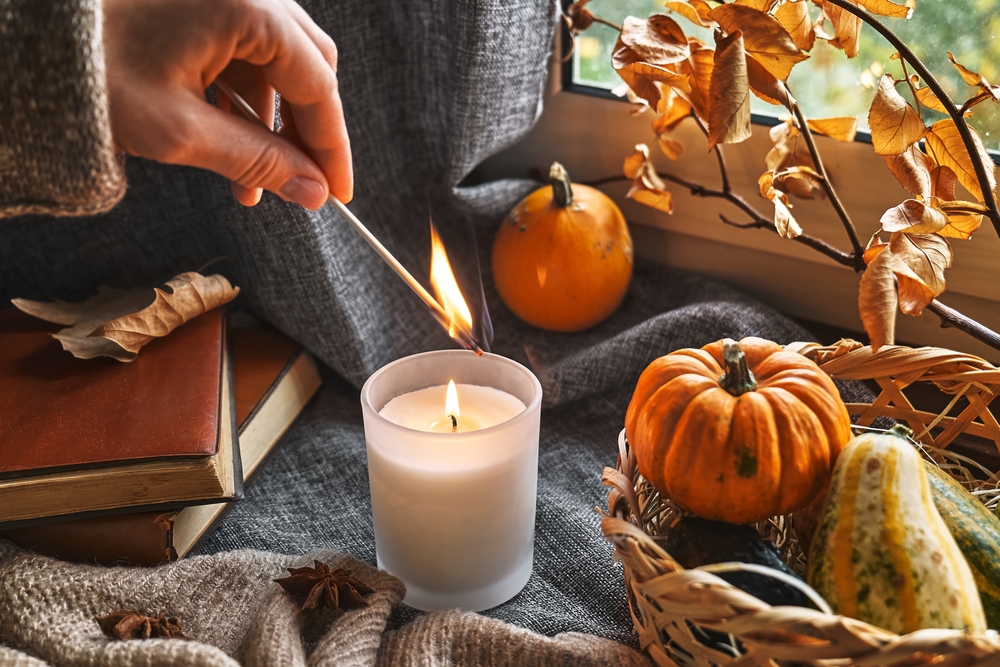 Hand with burning match lighting a candle on the windowsill with cozy autumn still life with pumpkins, knitted woolen sweater and books. Autumn home decor.