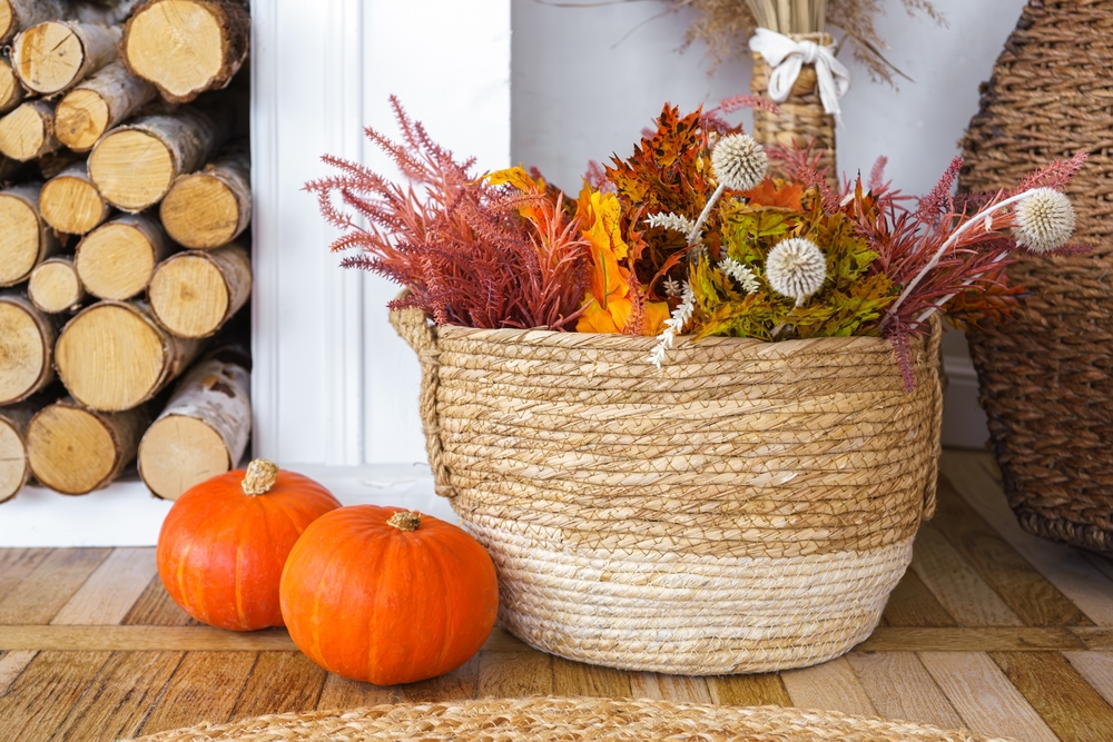 Woven basket brimming with vibrant dried flowers and plants, two small orange decorative pumpkins, set on wooden logs background. Seasonal home decor for fall holidays
