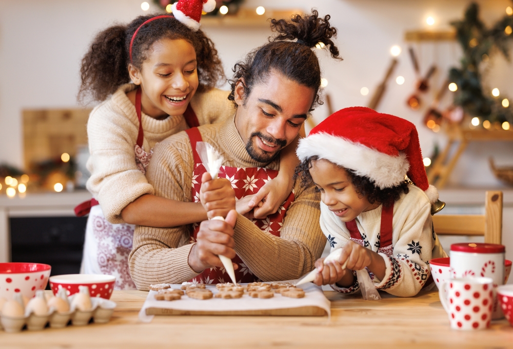 Happy african american family father and two kids in festive outfit making Christmas cookies together in kitchen at home during winter holidays, dad decorating xmas gingerbreads with small children
