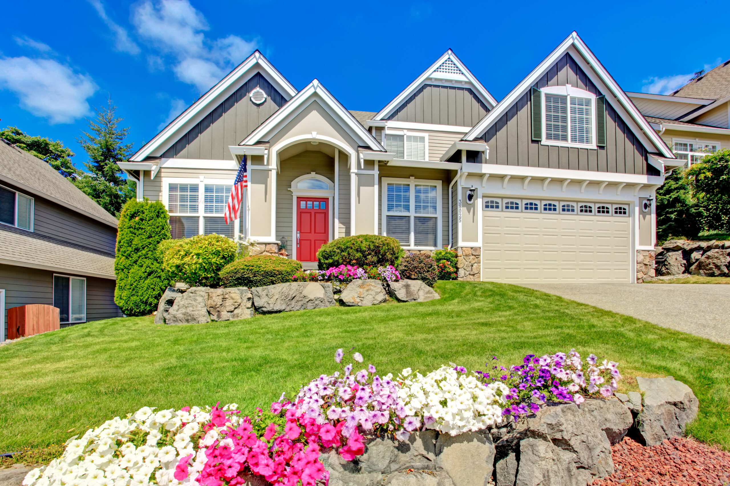 Grey house exterior with entrance porch and red door. Beautiful front yard landscape with vivid flower and stones