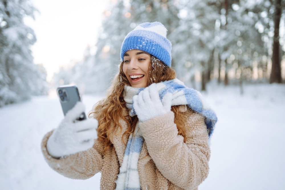 Happy woman in warm clothes making selfie with smartphone in winter forest against snow-covered trees and falling snow.
