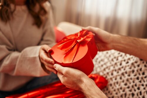 Happy Valentine's Day. Young couple in love exchanging gifts while sitting on the sofa in the living room at home. A man giving a heart-shaped gift box to his beloved woman.