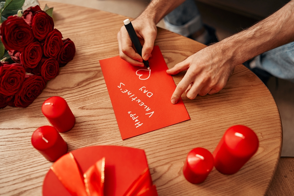 Happy Valentine's Day. A man writes a greeting card for his soulmate, there are also candles, a gift and a bouquet of roses on the table nearby.