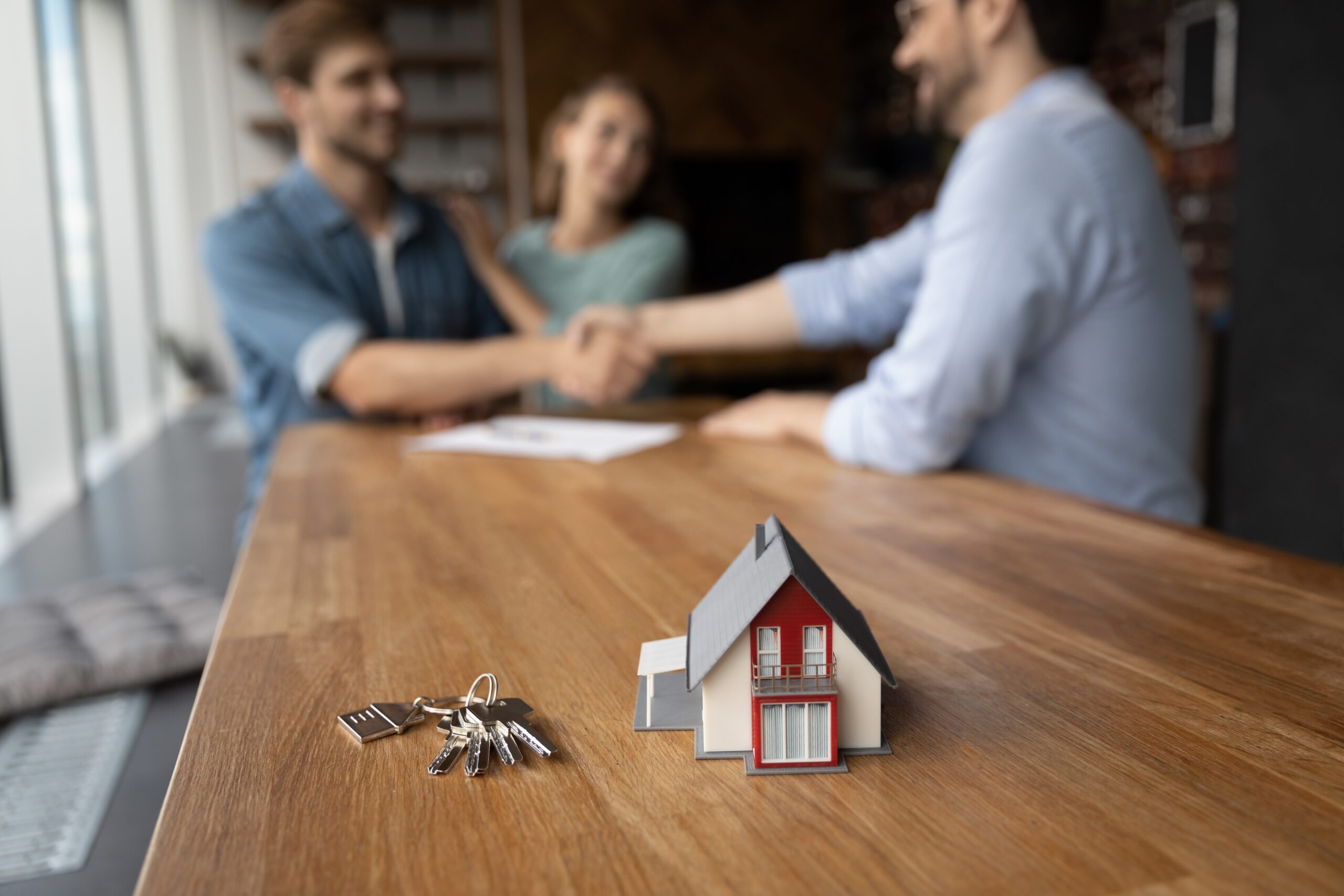 Toy house and keys with family couple of clients and real estate agent giving handshakes in background. Customers meeting with broker, property seller, lawyer, shaking hands over buying contract
